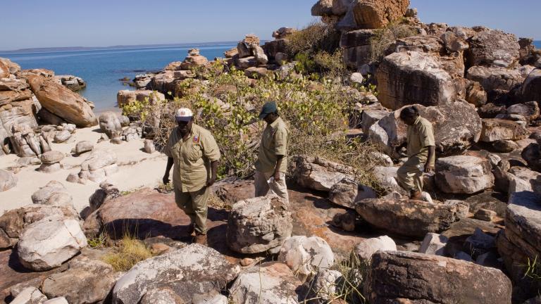 Three Indigenous rangers walking across rocks near the ocean
